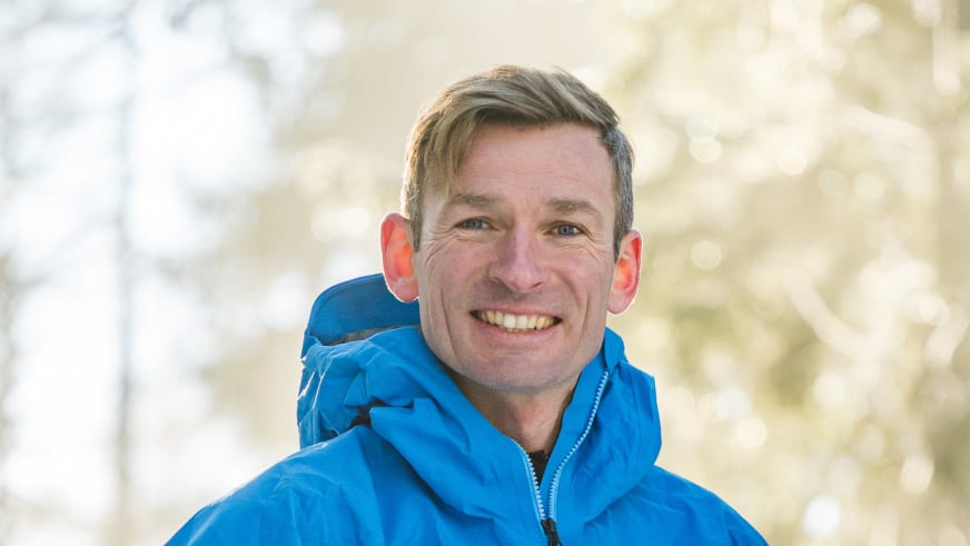 Bergführer Martin Riener lächelt in blauer Softshelljacke mit Kapuze vor verschwommener Berglandschaft ins Objektiv.