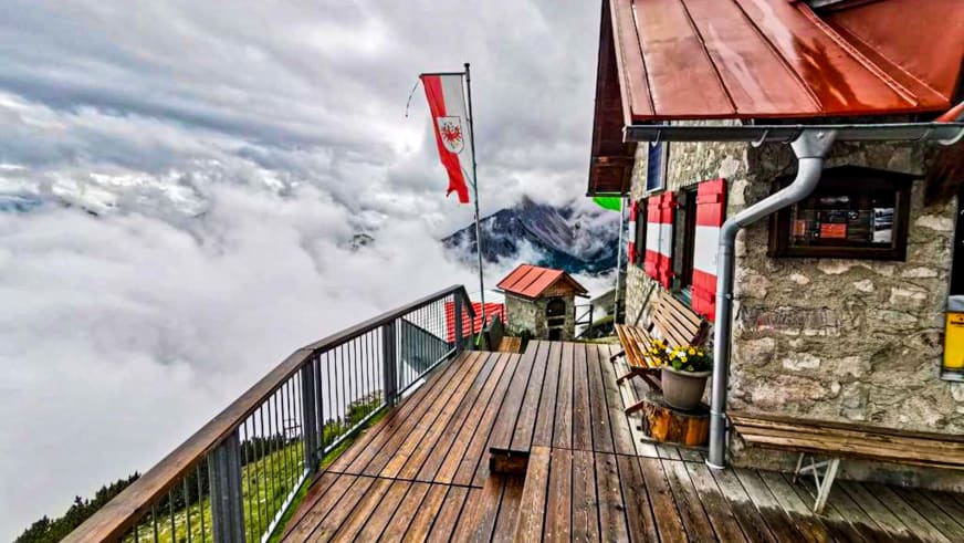 Holzterrasse der Bettelwurfhütte mit Blick über die Wolkendecke auf schneebedeckte Karwendel-Gipfel.
