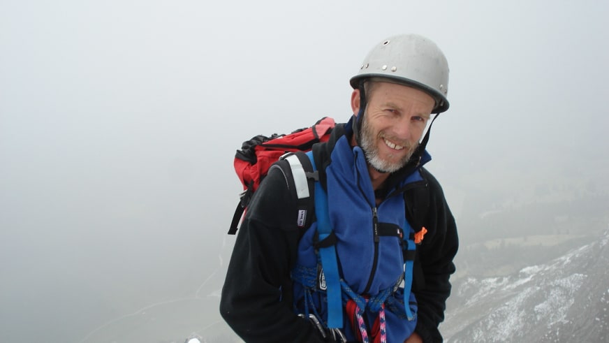 Bergwanderführer Helmut Meyer trägt grauen Kletterhelm und blaue Softshell-Jacke vor schneebedeckten Bergen.