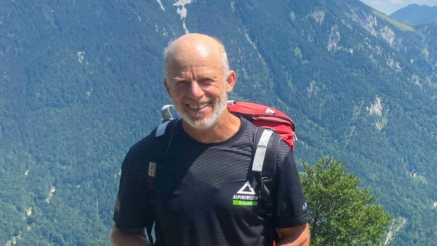 Bergwanderführer Helmut Meyer mit grauem Vollbart und schwarzem Rucksack lächelt vor grünen Bergen in die Kamera.