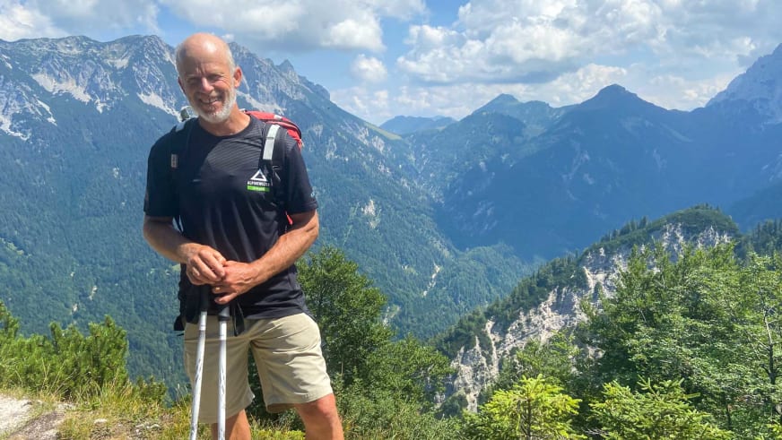 Bergwanderführer Helmut Meyer hält zwei Wanderstöcke und trägt einen Rucksack vor einem bewaldeten Bergtal.