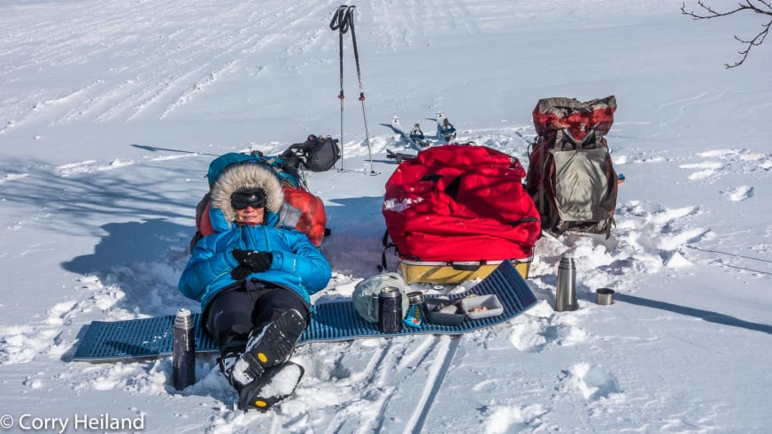 Bergwanderführerin Corry Heiland in blauer Daunenjacke mit Fellkapuze sitzt auf einer blauen Isomatte im Schnee und pausiert während ihrer Wintertour im verschneiten Fjell.