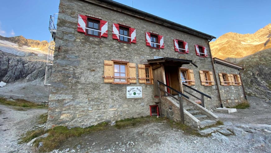 Außenansicht der Richterhütte aus Naturstein mit rot-weißen Fensterläden vor Bergkulisse.