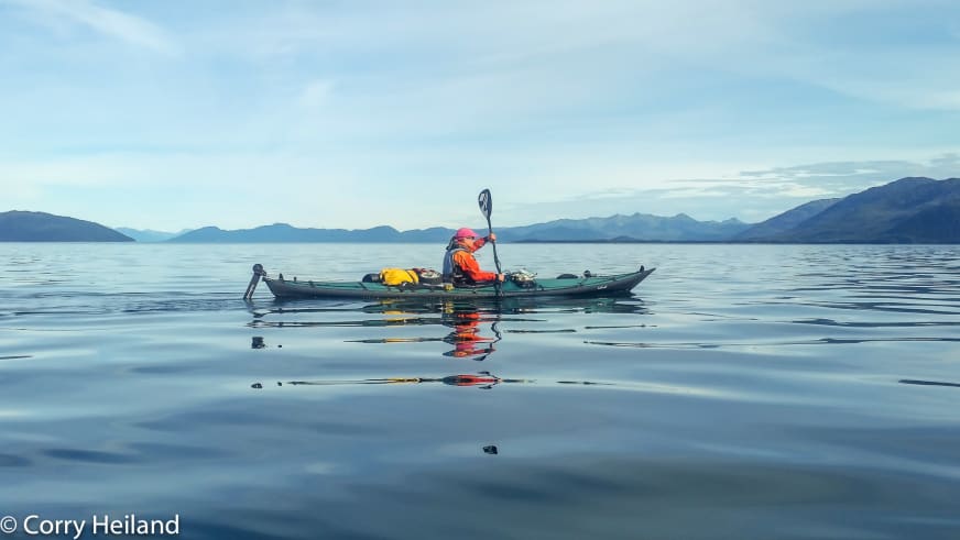 Im grünen Seekajak paddelt Bergwanderführerin Corry Heiland mit roter Schwimmweste auf spiegelglattem Wasser vor Gebirgskette.