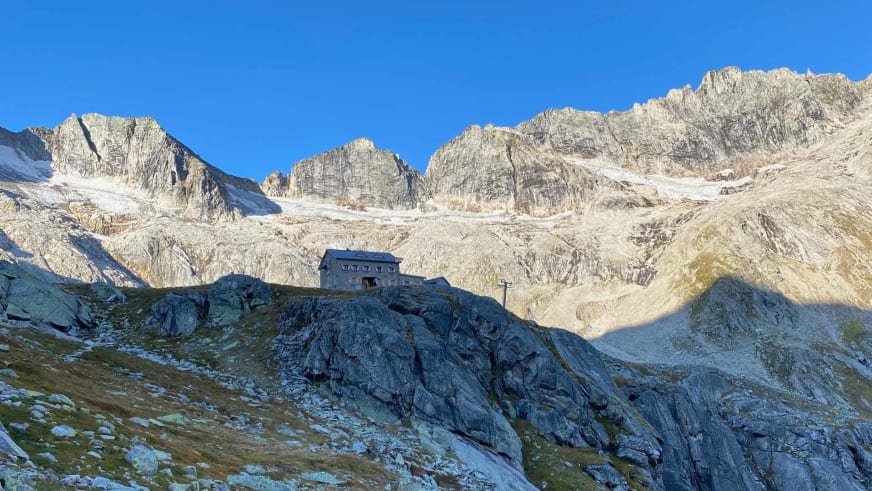 Die Richterhütte steht vor steilen Felswänden und schneebedeckten Gipfeln bei blauem Himmel.
