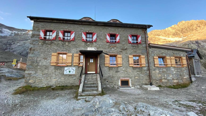 Die Richterhütte aus Naturstein mit rot-weißen Läden steht vor schneebedeckten Berggipfeln.