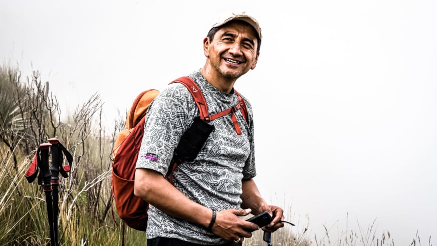 Mit beiger Basecap und orangefarbenem Rucksack hält Bergführer Marcelo Puruncajas ein elektronisches Gerät in Graslandschaft.