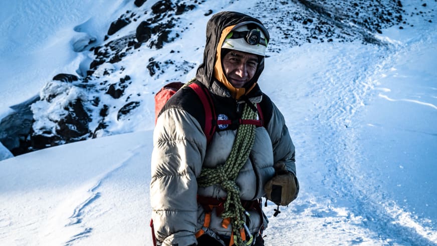 Bergführer Marcelo Puruncajas trägt helle Daunenjacke mit weißem Helm und grünem Kletterseil in schneebedeckter Berglandschaft.