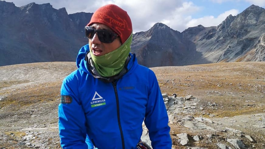Bergführer Marcelo Puruncajas trägt blaue Softshelljacke mit Alpine Welten Logo, rote Mütze und Sonnenbrille in karger Hochgebirgslandschaft.