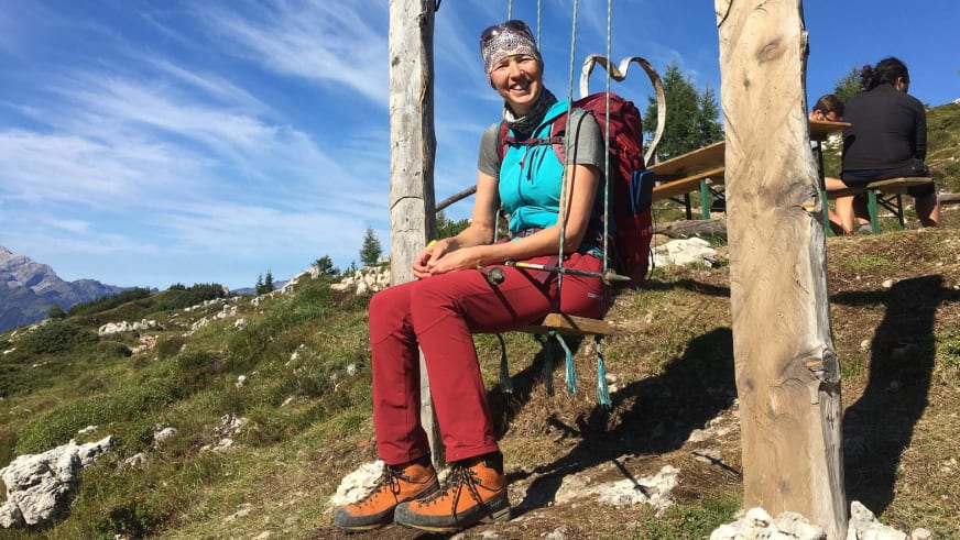 Bergwanderführerin Ruth Zeller sitzt lächelnd auf einer Holzbank am Rastplatz einer Bergwiese mit rotem Wanderrucksack.