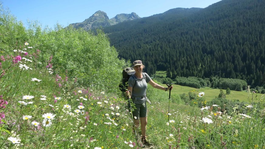 Auf einer blühenden Almwiese mit weißen Margeriten steht Bergwanderführerin Ruth Zeller mit Wanderstöcken und großem Rucksack.