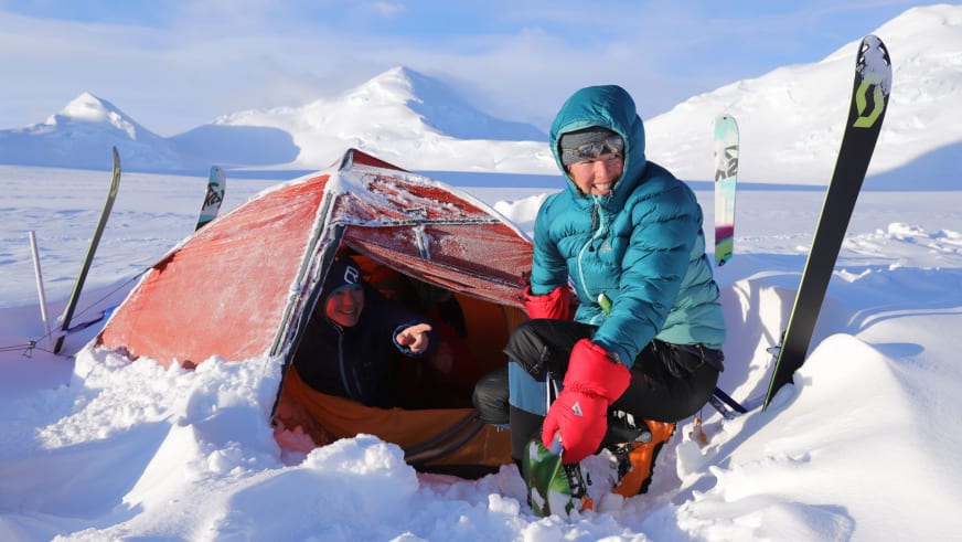 Im Schnee vor einem rot-orangen Zelt sitzt Bergwanderführerin Ruth Zeller in türkisfarbener Daunenjacke mit aufgestellten Skiern ringsum.