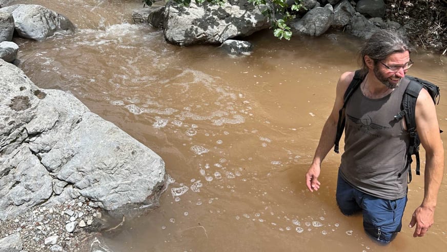 Bergführer Thomas Scherzer steht mit braunem Tanktop und Rucksack lachend in schlammigem Wasser bis zur Hüfte.