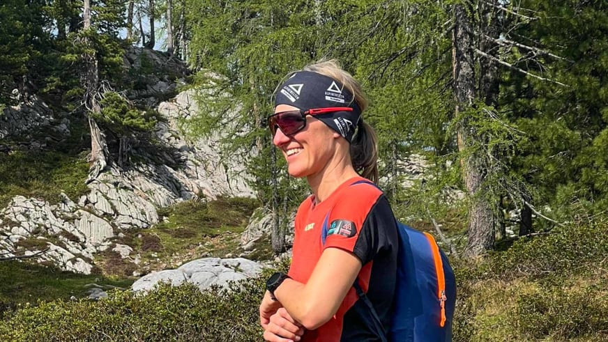 Bergwanderführerin Elisabeth Götzinger mit roter Sonnenbrille und orangem Shirt vor Kalkstein-Berglandschaft.