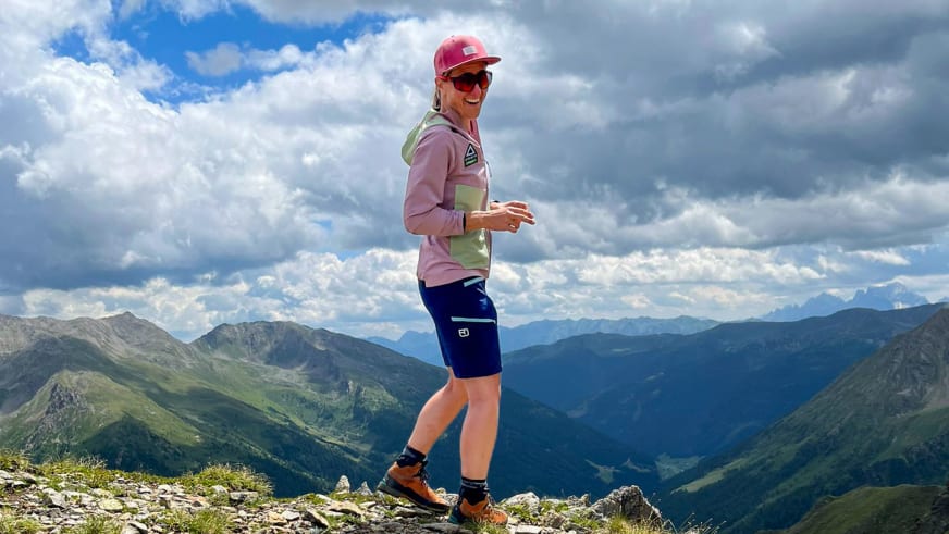Bergwanderführerin Elisabeth Götzinger läuft mit roter Kappe und gelbem Rucksack über steinigen Berggrat mit Alpenpanorama.