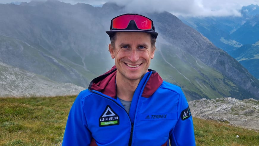 Portrait von Bergführer Christian Färber mit schwarzer Kappe und blauer Alpine Welten Jacke auf grüner Bergwiese.