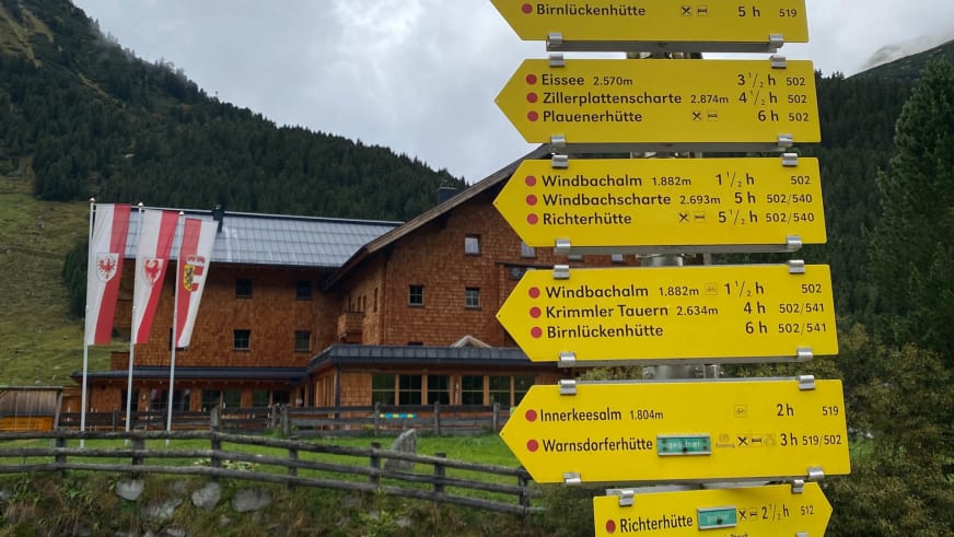 Gelber Wegweiser mit Wanderzielen vor dem Krimmler Tauernhaus und österreichischen Fahnen.