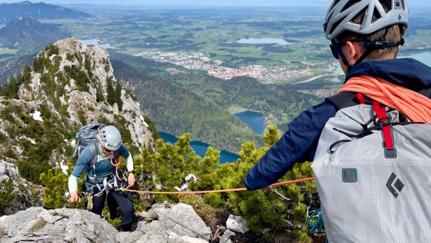 Bergführer Christian Färber klettert mit zweiter Person am orangen Seil über steilen Felsgrat mit Panoramablick auf blauen See im Tal.