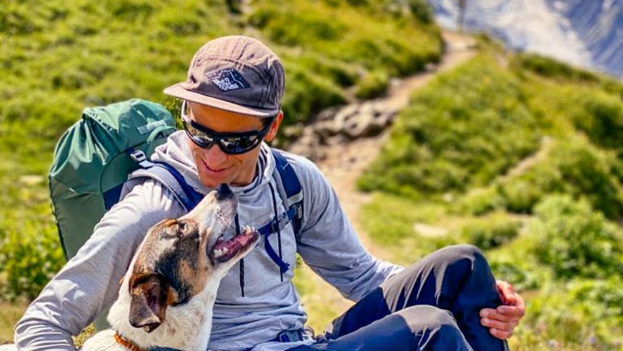 Bergführer Christian Färber sitzt entspannt auf Wiese und streichelt hellen Hund mit orangem Halsband.