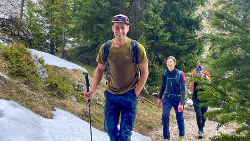 Bergführer Christian Färber wandert mit roter Kappe und blauem Rucksack auf Bergweg durch Nadelwald, gefolgt von zwei Personen.