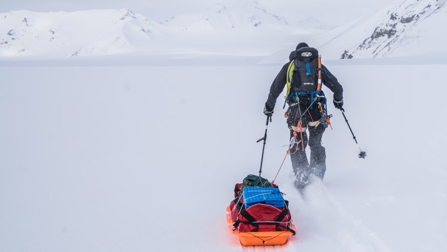 Bergwanderführer Jonas Haass in schwarzer Winterkleidung zieht beladenen rot-blauen Transportschlitten durch tiefen Schnee.