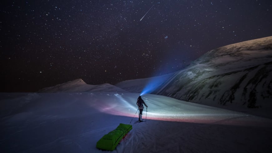 Nächtliche Szene zeigt Bergwanderführer Jonas Haass mit Stirnlampe auf Skiern unter Sternenhimmel mit Sternschnuppe.