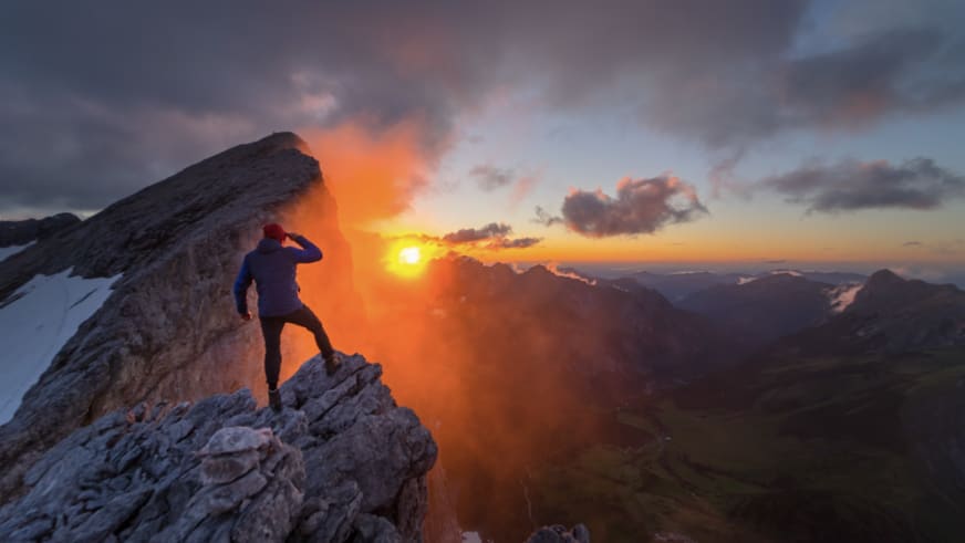 Bergwanderführer Jonas Haass in dunkler Kleidung und roter Mütze steht auf schmalem Felsgrat vor orange-gelbem Sonnenaufgang-Himmel.