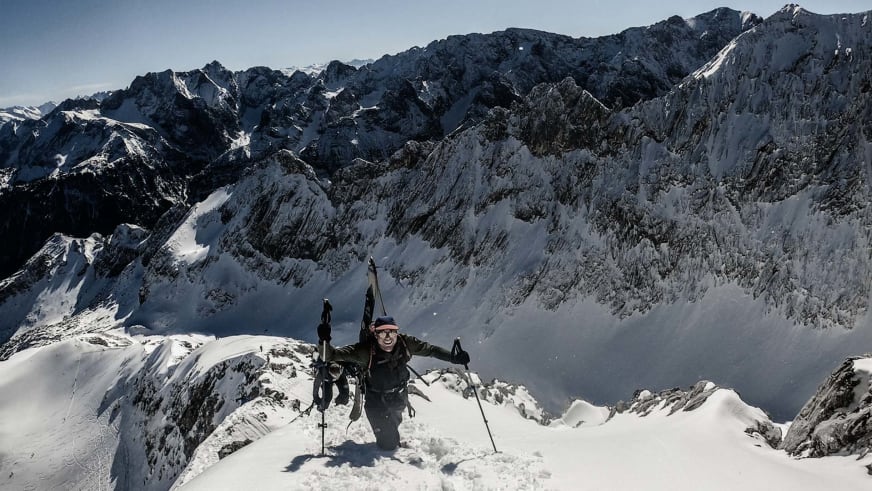 Bergwanderführer Jonas Haass steht mit Skibrille und roter Mütze auf verschneitem Gipfel vor Hochgebirgspanorama.