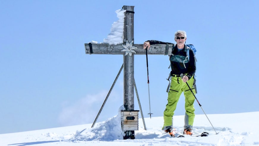 Am Gipfelkreuz mit der Zahl 92 steht Bergführer Hermann Böhler mit Tourenskiern bei strahlendem Sonnenschein.