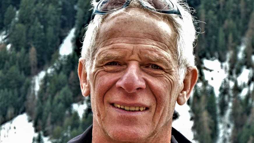 Im Winter zeigt ein Portrait Bergführer Hermann Böhler mit Skibrille auf der Stirn vor verschneiten Nadelbäumen.