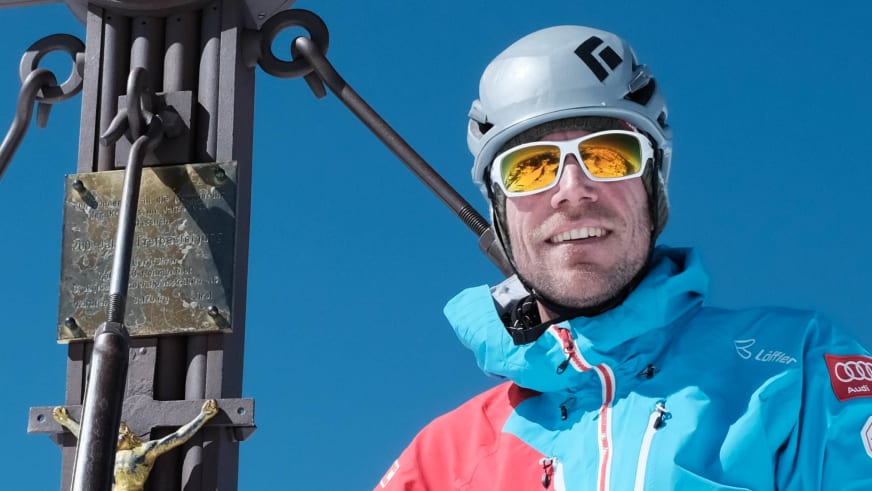 Bergführer Michael Eberharter lächelt in die Kamera, trägt weißen Helm und türkis-blaue Skijacke am Gipfel.