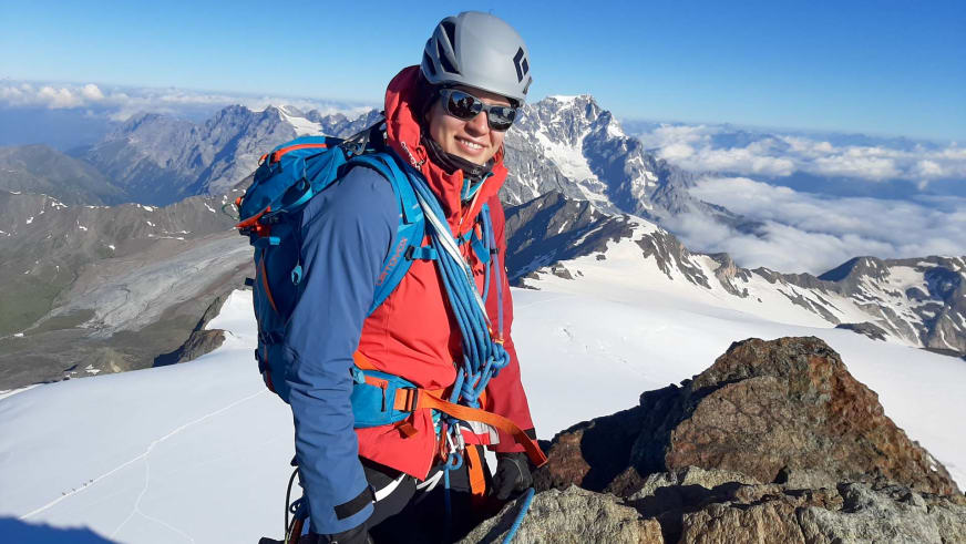 Bergwanderführerin Tatjana Reichert steht mit weißem Helm und roter Jacke auf verschneitem Gipfel vor Hochgebirgspanorama.