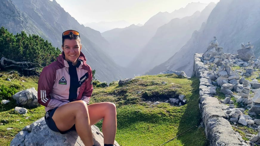 Auf einem Felsblock sitzt Bergwanderführerin Tatjana Reichert in rosa Outdoorjacke vor grüner Almlandschaft.