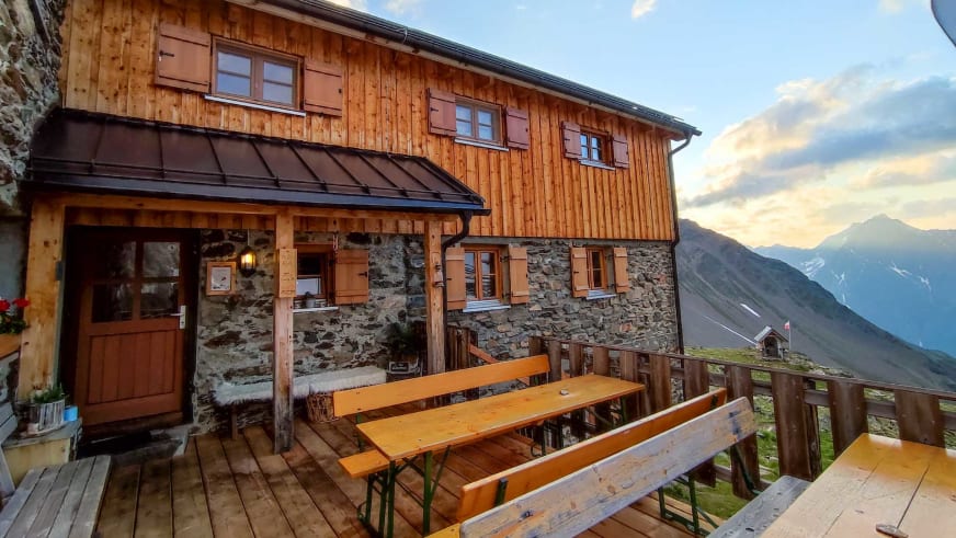 Warme Abendbeleuchtung der Kaunergrathütte mit Holzterrasse und Bergketten im Hintergrund.