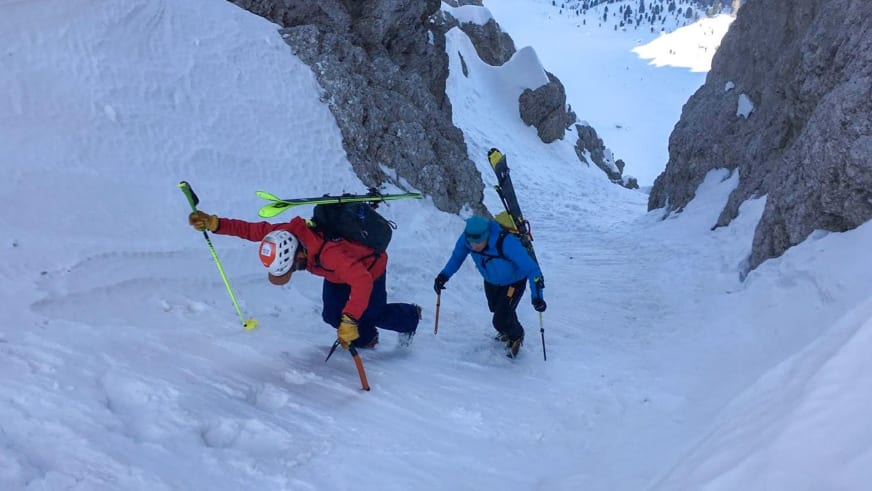 Durch steiles verschneites Couloir steigen zwei Skitourengeher auf, geführt von Bergführer Tommaso Cappelletto in roter Jacke.