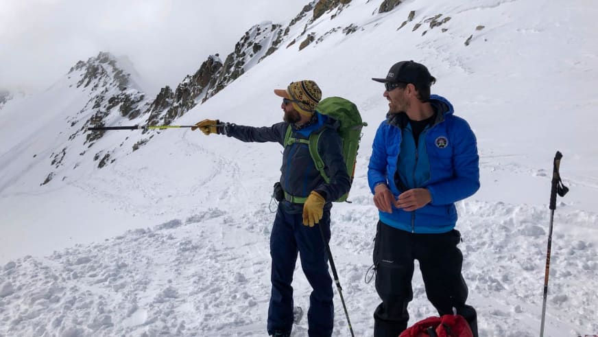 Im verschneiten Hochgebirge pausieren zwei Personen, eine davon zeigt mit Skitourenstock zu den Bergen, begleitet von Bergführer Tommaso Cappelletto.