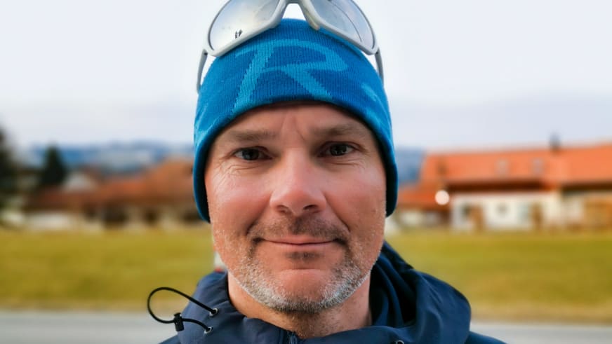 Portrait von Bergführer Tadej Zorman mit blauer Strickmütze und dunkler Funktionsjacke vor bewaldeten Hügeln und Gebäuden mit orangenen Dächern.