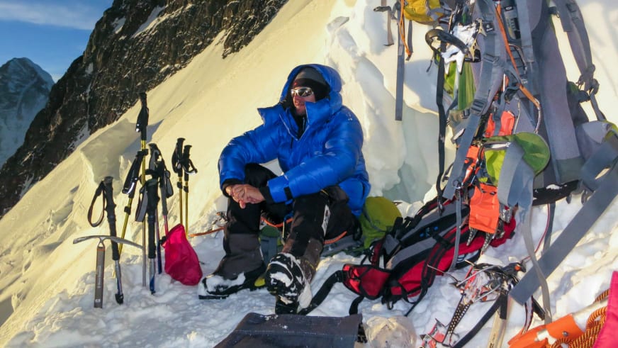 Bergführer Tadej Zorman sitzt in blauer Daunenjacke im Schnee und pausiert umgeben von umfangreicher Hochtourenausrüstung mit Pickeln und Seilen.