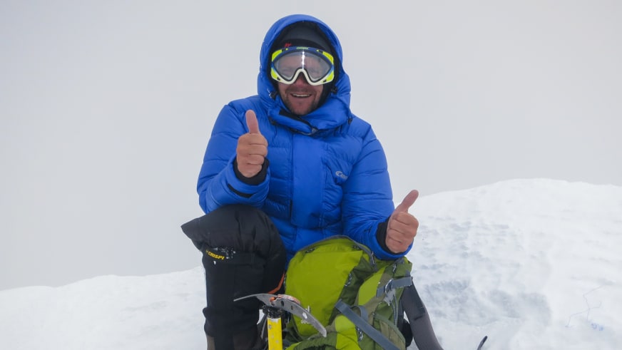 Bergführer Tadej Zorman sitzt im Schnee mit blauer Daunenjacke und zeigt Daumen hoch, vor ihm liegt ein grüner Rucksack mit gelbem Eispickel.