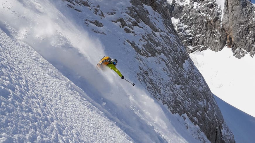 Bergführer Tadej Zorman fährt in gelber und grüner Ski-Kleidung einen steilen, verschneiten Hang hinunter und wirbelt Schneewolken auf.