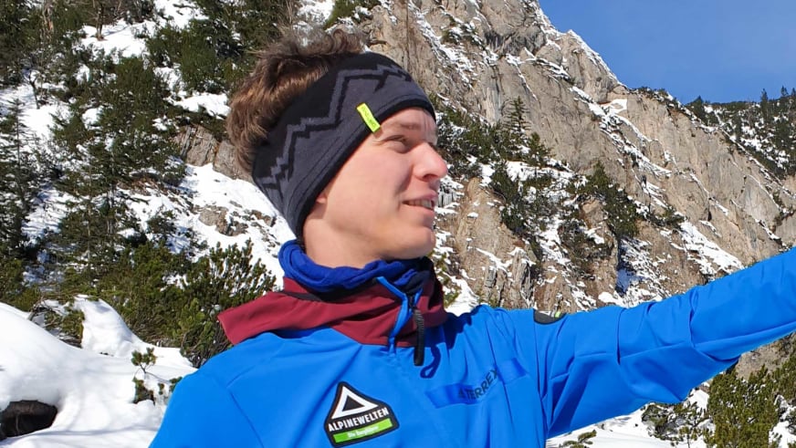 Bergwanderführer Uwe Roth macht ein Selfie vor verschneiten Felswänden und trägt blaue Outdoorjacke mit Alpine Welten Logo.