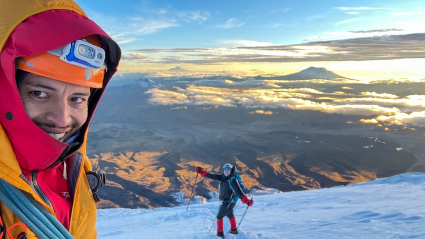 Bei Sonnenaufgang lächelt Bergführer José Salazar mit roter Kapuze in die Kamera, während ein Skitourengeher im Hintergrund aufsteigt.