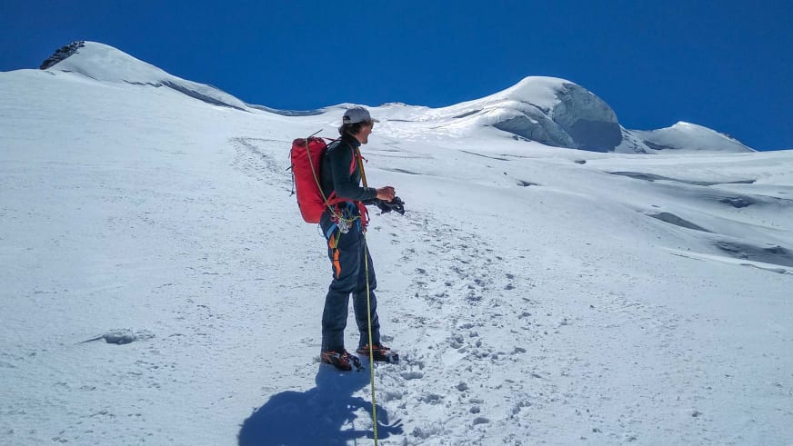 Auf einem weiten Firnfeld steht Bergführer Davide Marinovich mit rotem Rucksack und Steigeisen vor schneebedeckten Berggipfeln.