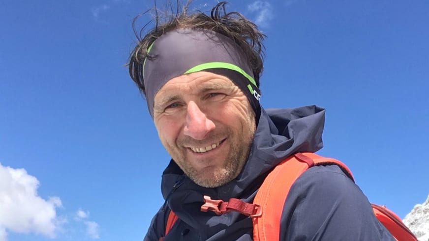 Lächelnder Bergführer Davide Marinovich trägt grünes Stirnband, graue Softshelljacke und orangefarbenen Rucksack vor blauem Himmel.