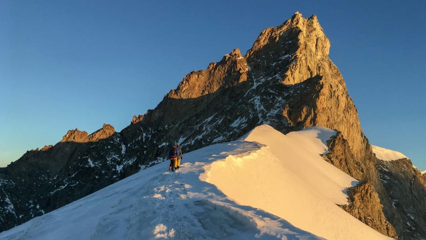 Zwei Bergsteiger in Seilschaft bewegen sich auf schmalem Firnkamm aufwärts, Bergführer Davide Marinovich führt im Morgenlicht zum Felsgipfel.