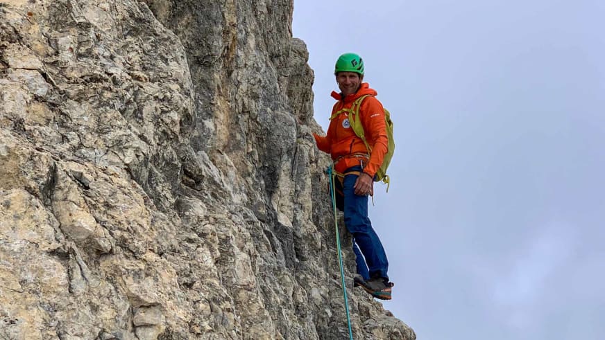 An einer steilen Felswand aus hellem Kalkstein steht Bergführer Davide Marinovich in orangener Daunenjacke mit grünem Helm und Klettergurt.
