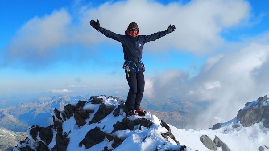 Bergführer Davide Marinovich steht mit ausgebreiteten Armen auf einem verschneiten Felsgipfel mit Klettergurt und bunten Karabinern.