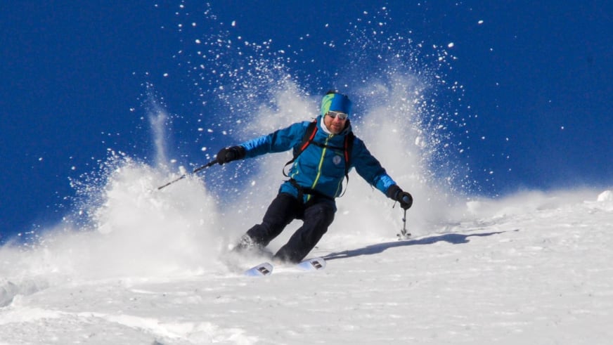 Bergführer Davide Marinovich fährt in blauer Winterjacke durch tiefen Pulverschnee und erzeugt große Schneefontänen zu beiden Seiten.