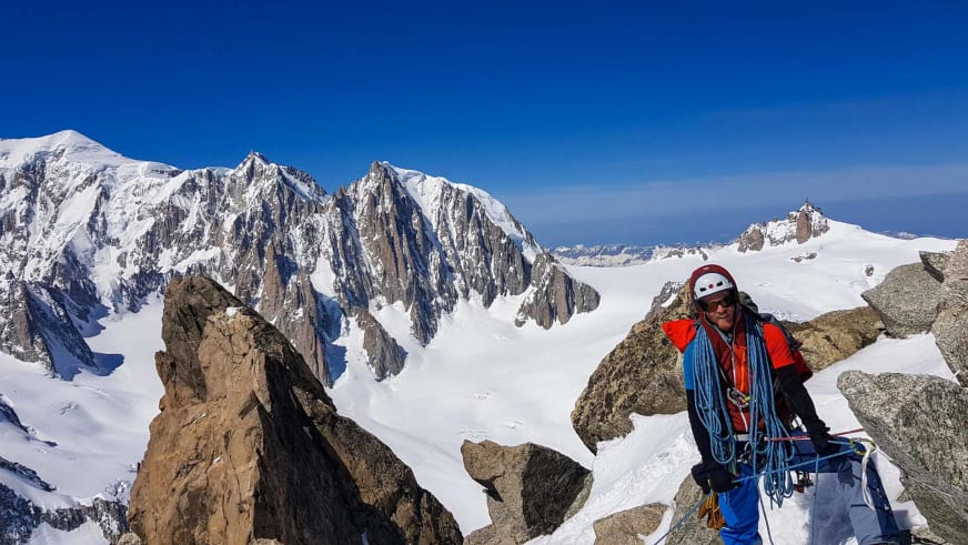 Auf einem schneebedeckten Felsvorsprung steht Bergwanderführer Christoph Knappe in roter Bergbekleidung mit weißem Helm und blauem Seil.