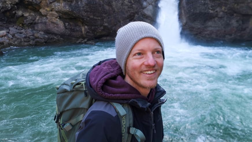 Bergwanderführer Christoph Knappe mit grauer Strickmütze und violetter Daunenjacke vor türkisfarbenem Gebirgsbach mit Wasserfall.